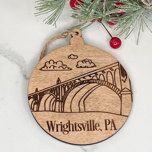 A wooden ornament in the shape of a circle with an etched image of a bridge and the text 'Wrightsville, PA'. It is hanging by a string, displayed against a marble background with Christmas decorations.